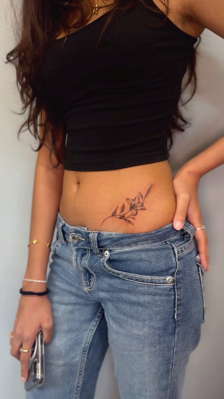 Floral tattoo on a person's lower abdomen wearing a black top and jeans.