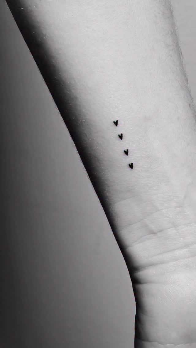 Four small black heart tattoos in a vertical line on a forearm.