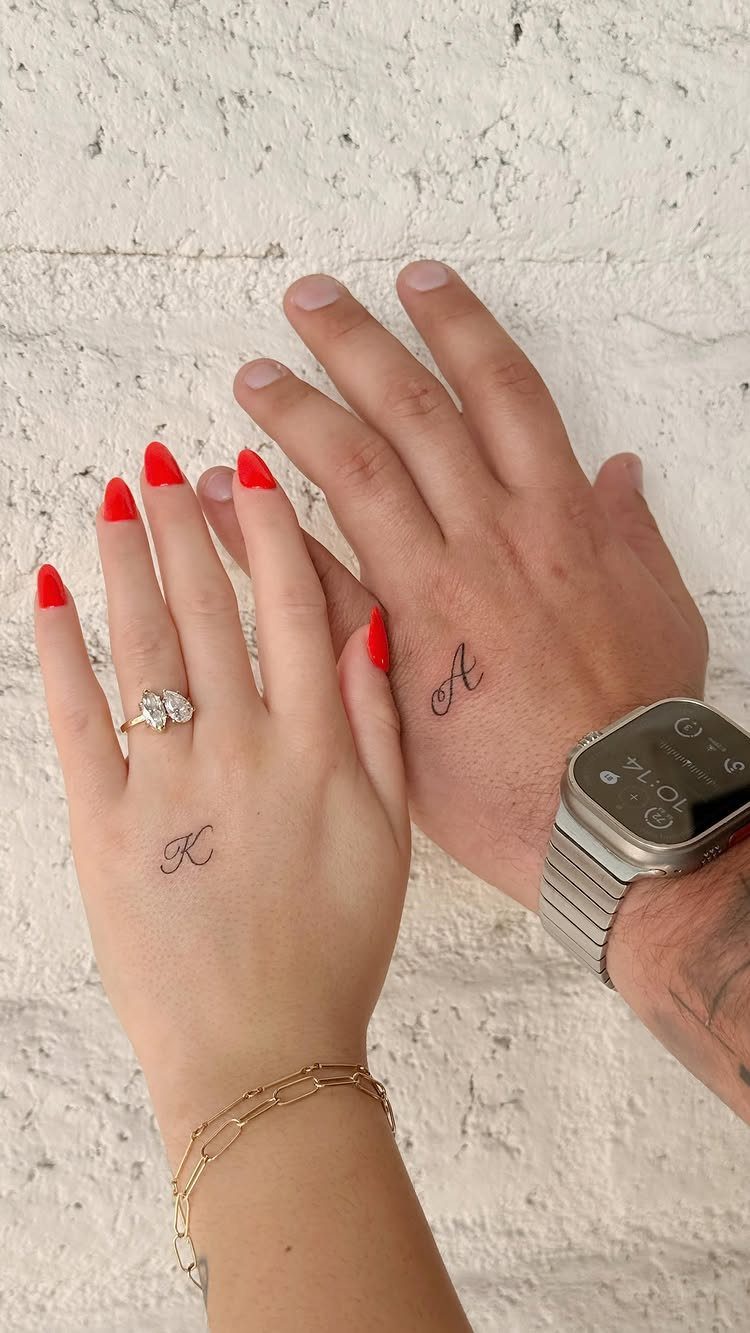 Two wrists with small cursive initial tattoos 'K' and 'A' against white wall.