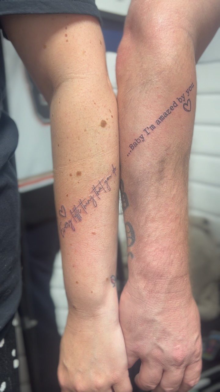 Two forearms with matching cursive text tattoos and small hearts in black ink.