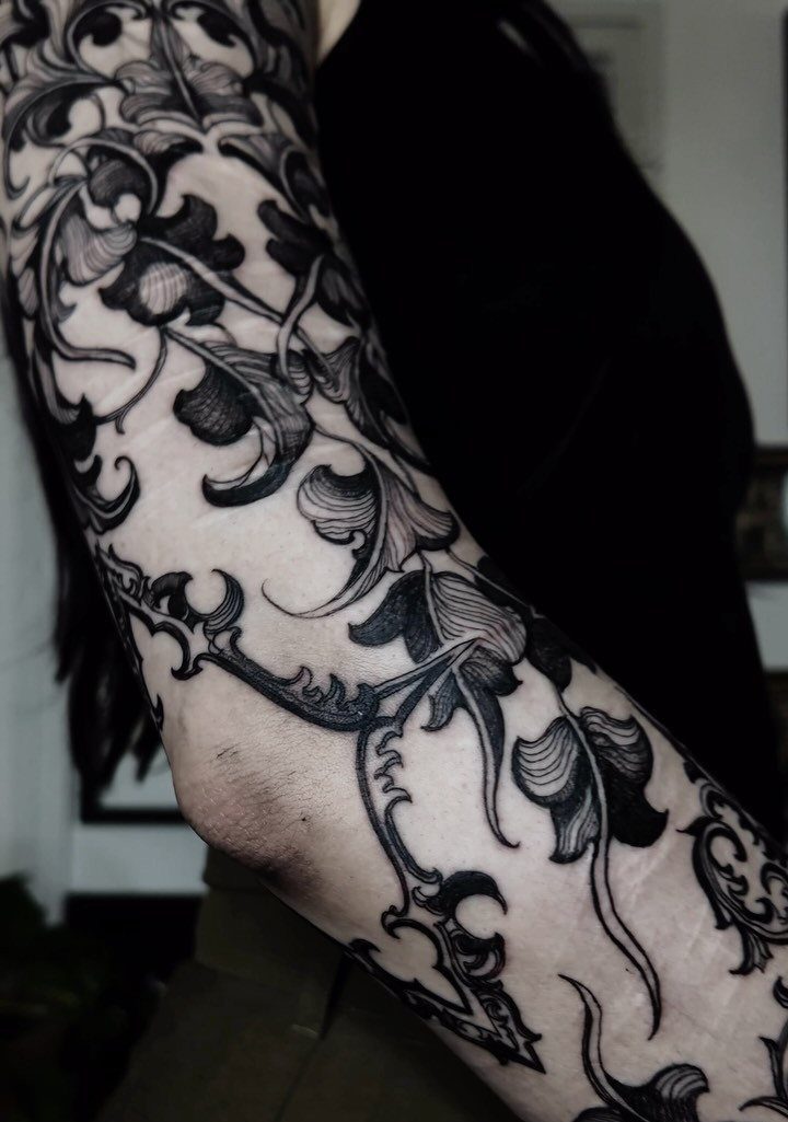 Ornate black ink baroque scrollwork tattoo covering a person's arm from shoulder to elbow.