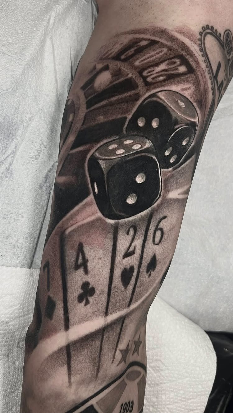 Bicep tattoo featuring black and grey dice, playing cards, and a roulette wheel.