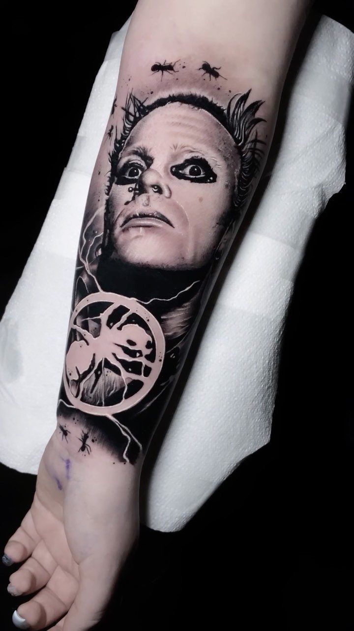 Forearm tattoo of a haunting face and circular symbol in black ink.