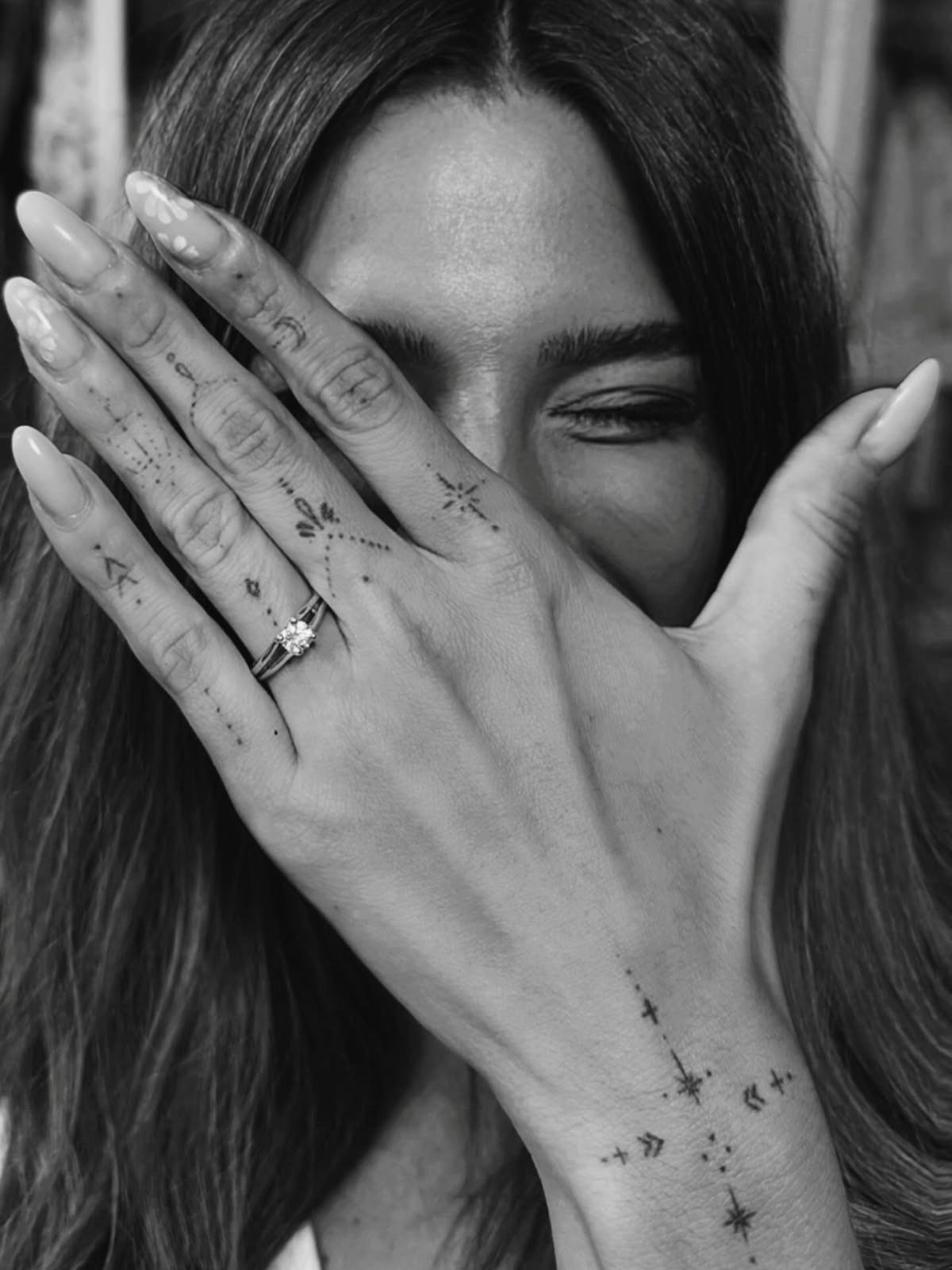 Woman's hand with small black symbol tattoos on fingers and wrist covering her face.