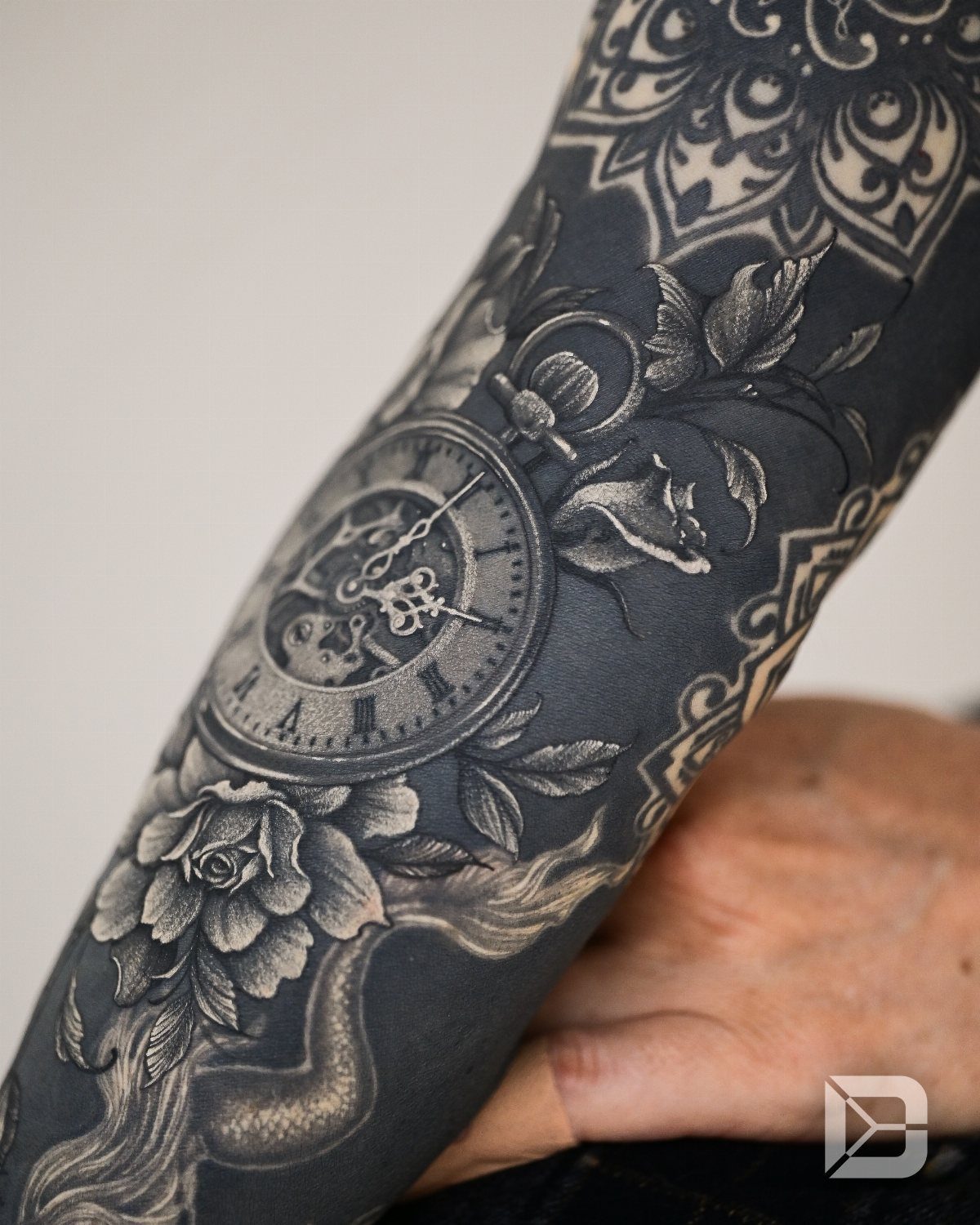 Black ink tattoo cover-up on an arm featuring a pocket watch and roses.