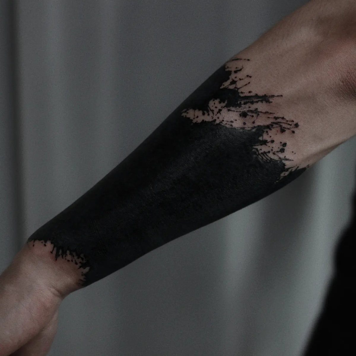 Forearm with a solid black ink tattoo cover up with splattered edges.