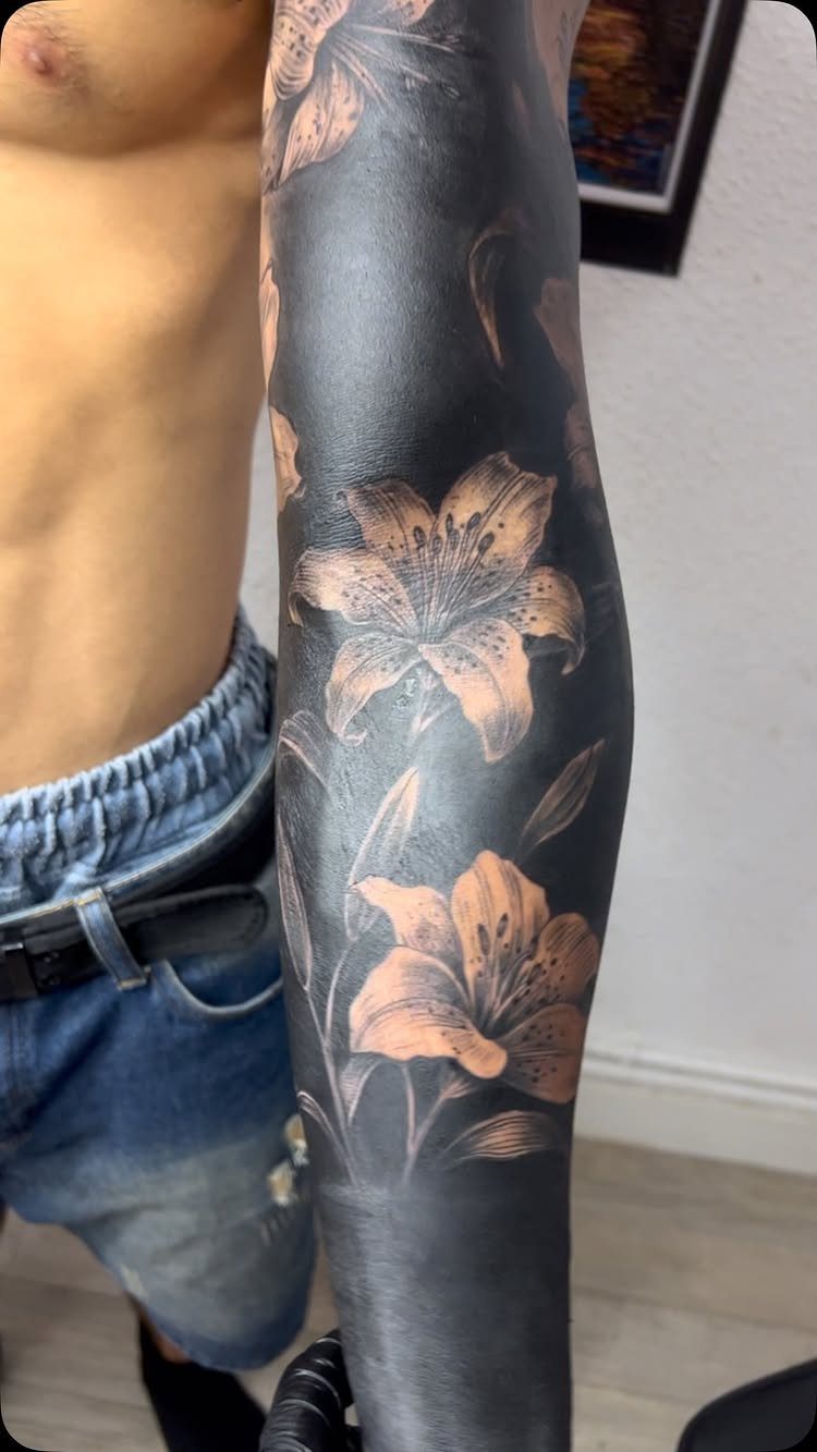 Arm with a blackout sleeve tattoo featuring light-colored lily flowers.