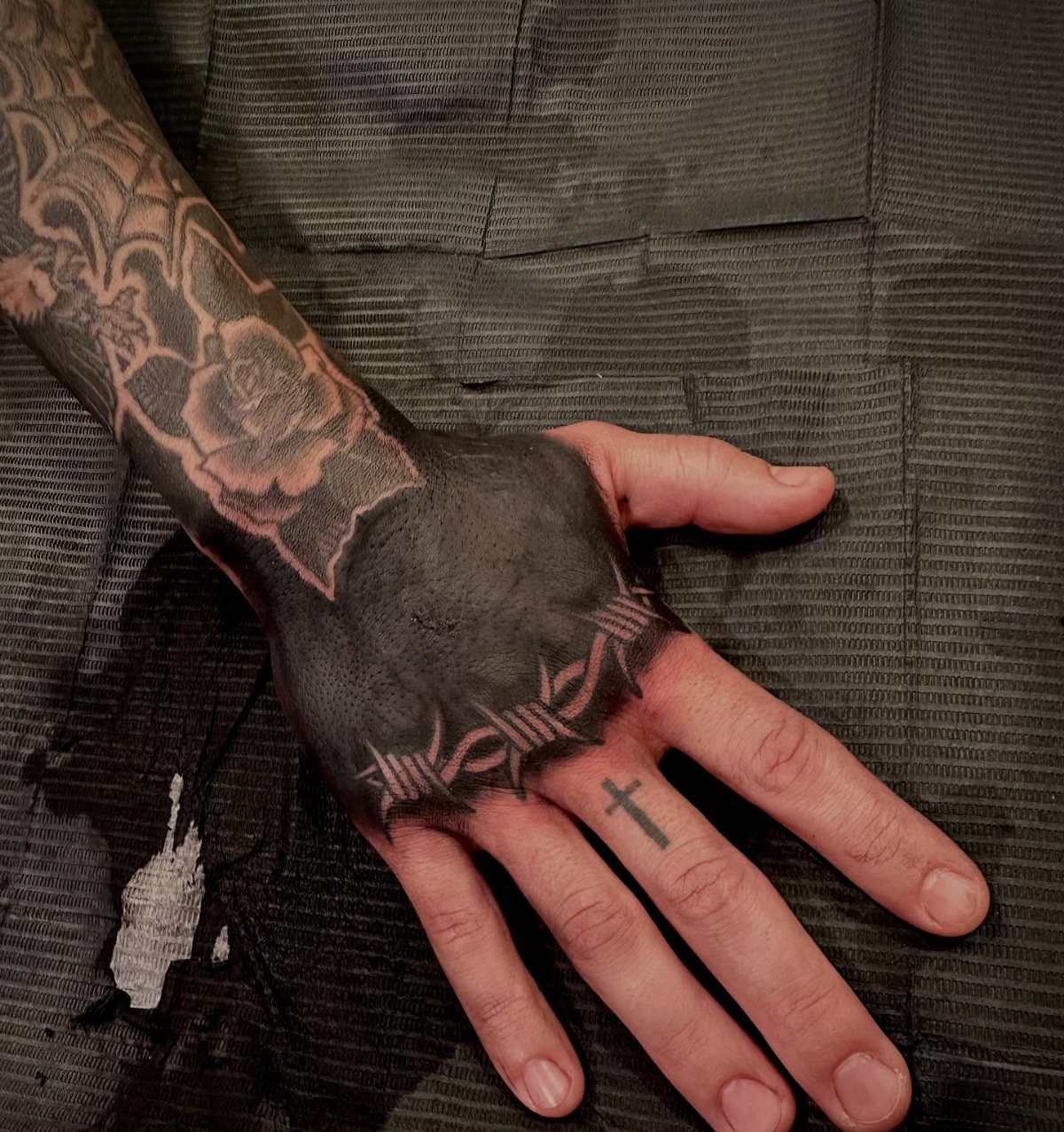 Hand with solid black ink tattoo featuring barbed wire and a small cross.