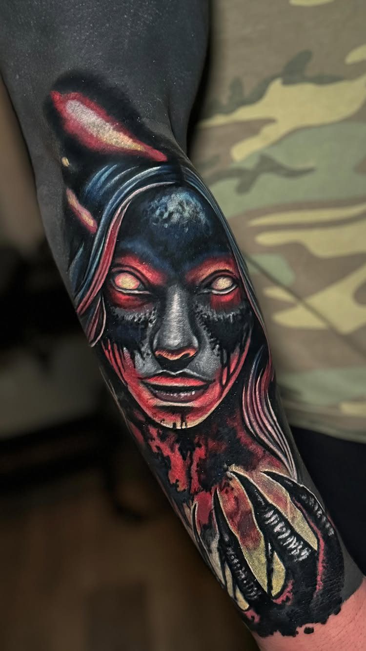 Demonic female face tattoo with glowing eyes on a blacked-out forearm.