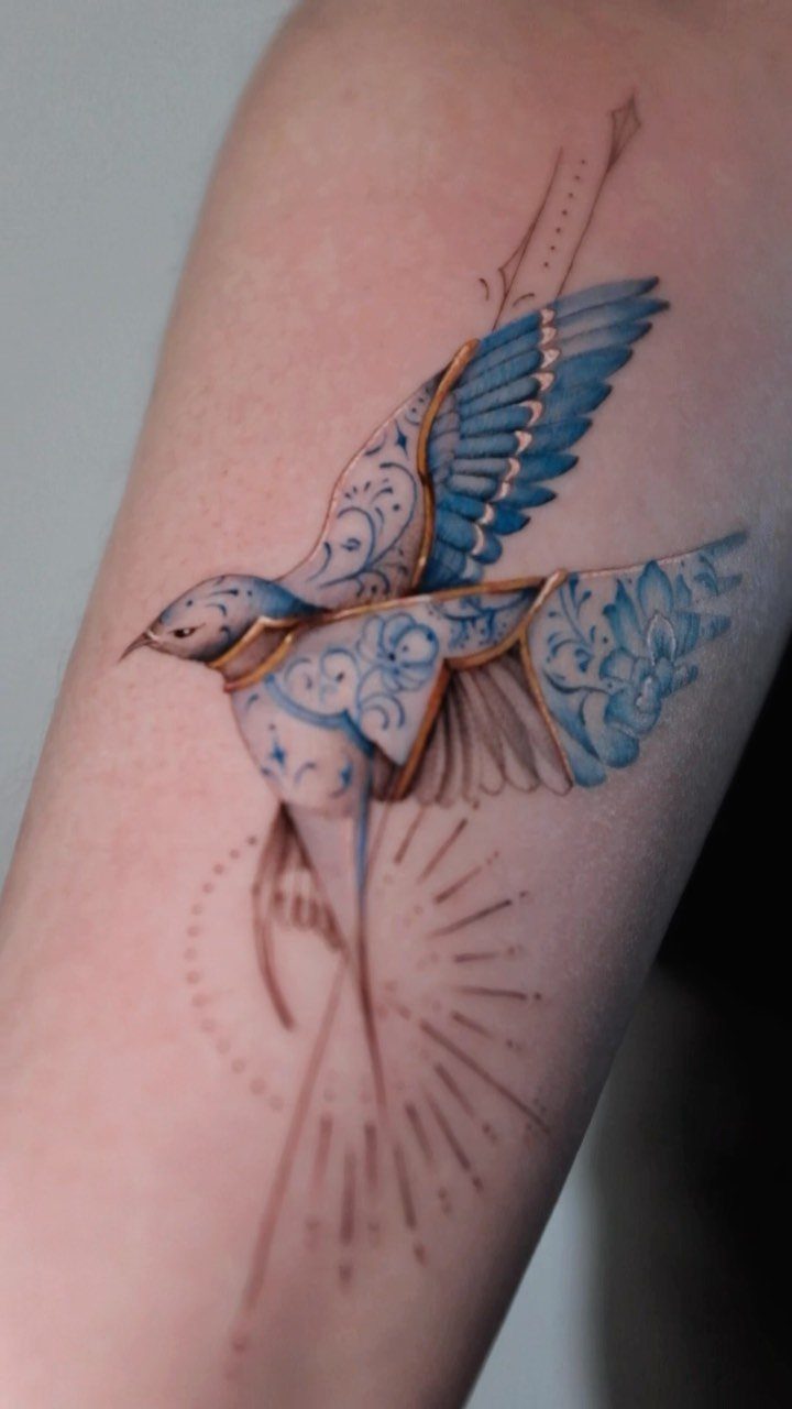 Blue bird tattoo with gold accents and floral patterns on a person's arm