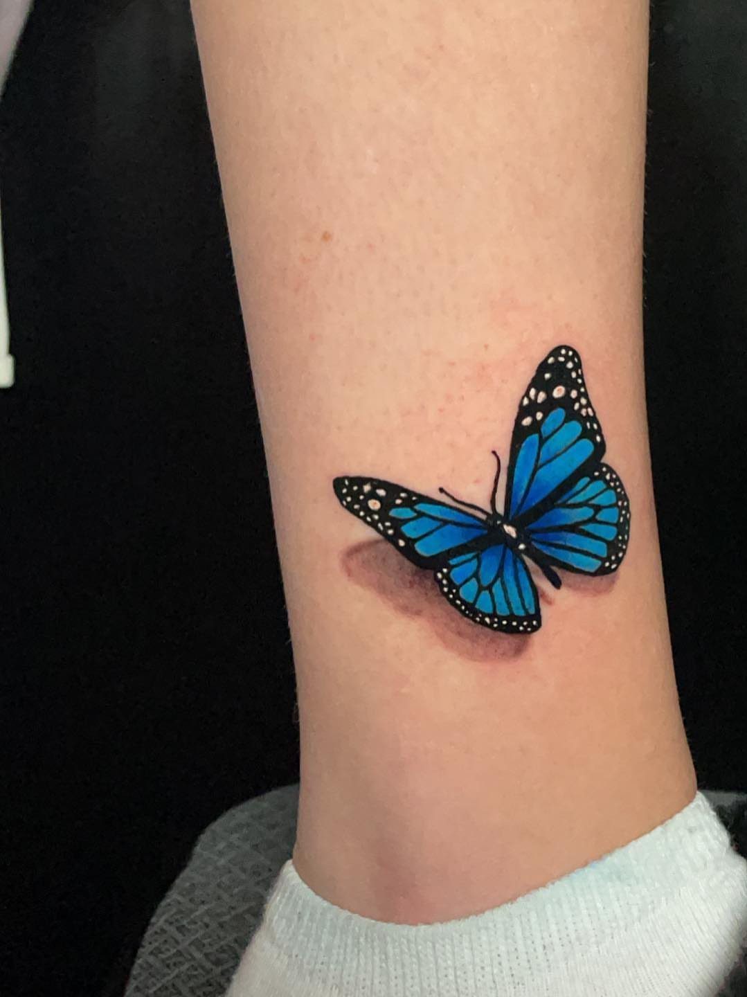 Blue butterfly tattoo on an ankle with detailed black wing edges and shading.