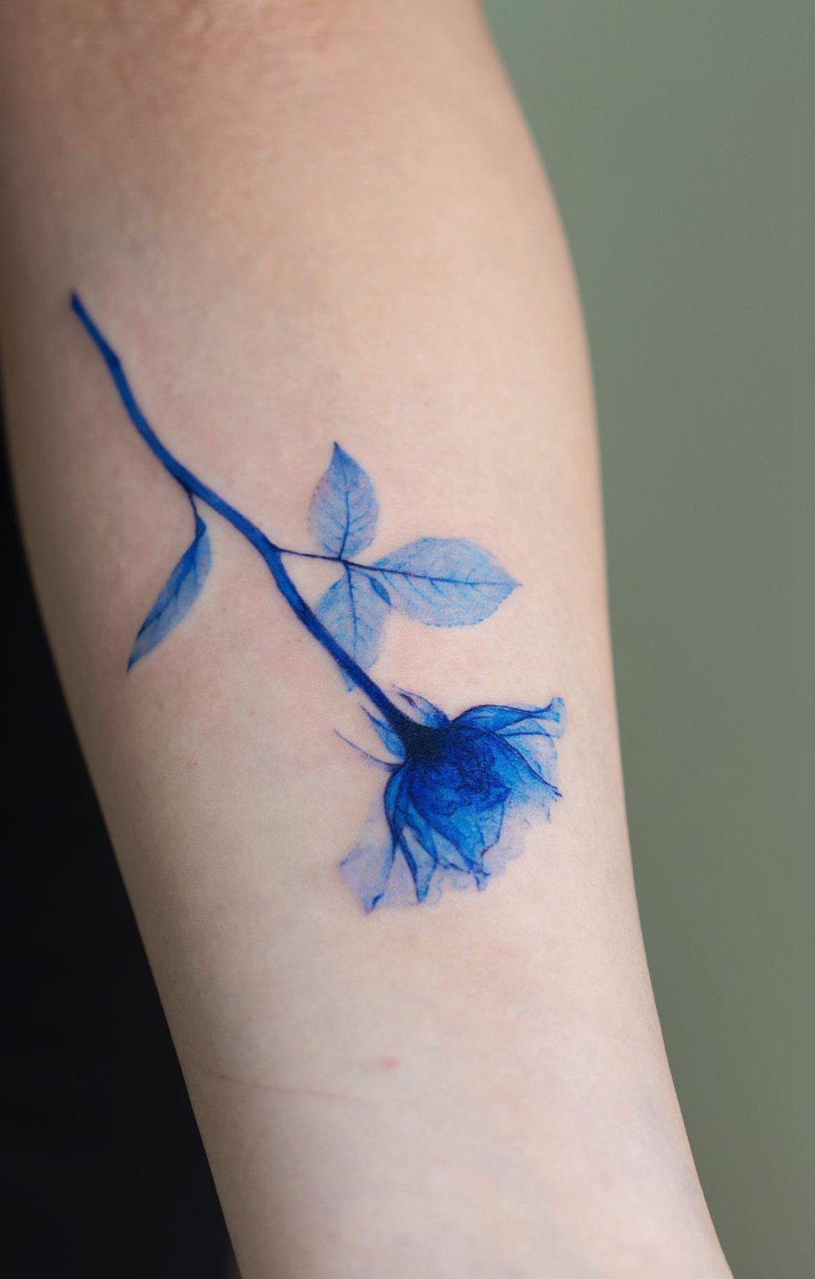 Blue ink tattoo of a rose with stem and leaves on a person's arm.