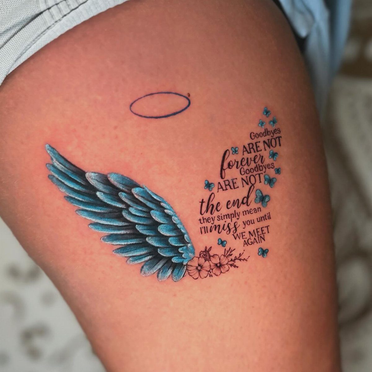 Blue ink tattoo of a wing and memorial text on a person's thigh
