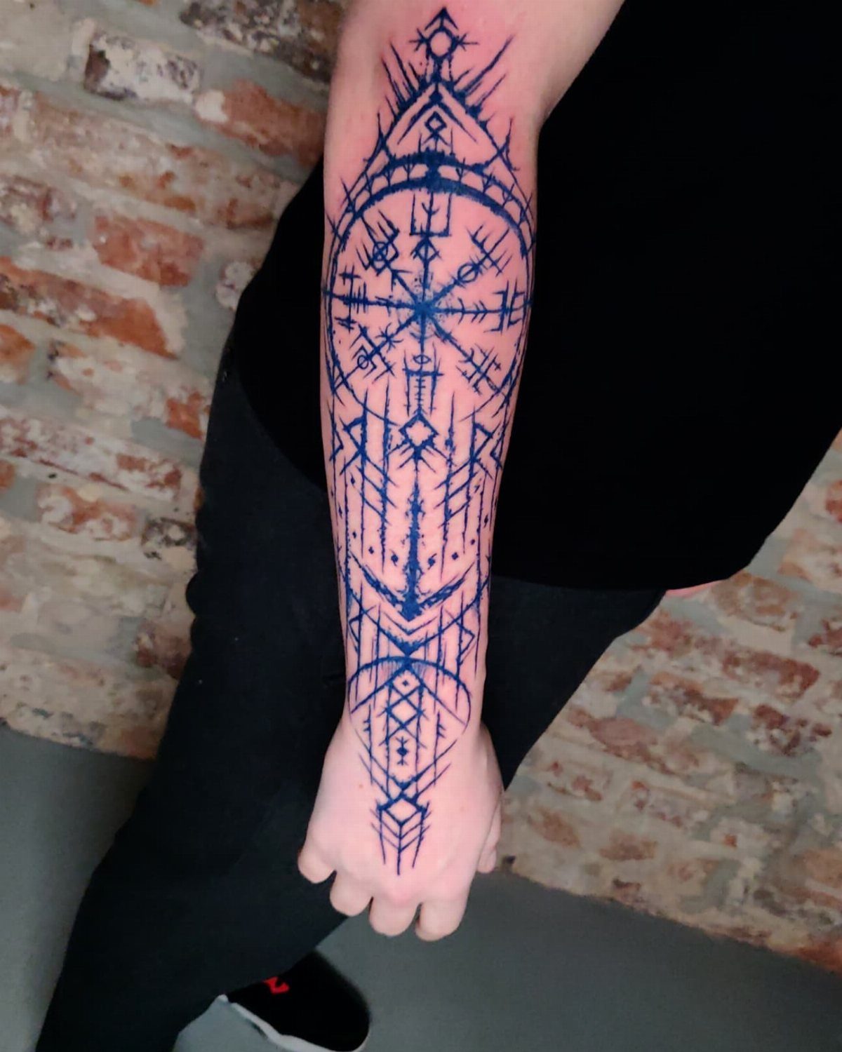 Intricate geometric blue ink tattoo on a person's forearm against a brick wall.