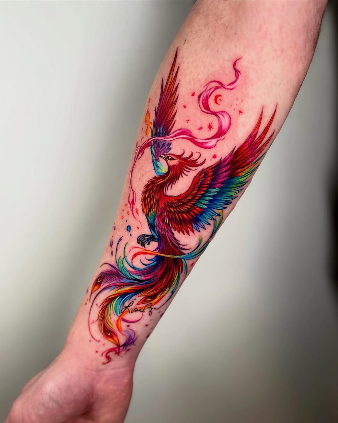 Vibrant multicolored phoenix tattoo with flowing feathers on a person's forearm.