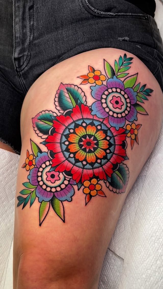 Colorful floral tattoo on a thigh featuring red, purple, and orange flowers with leaves.