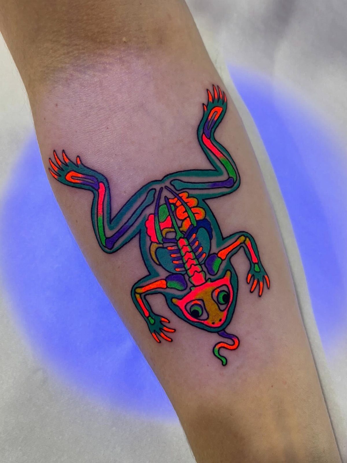 Neon frog skeleton tattoo with glowing orange and green ink on a forearm.