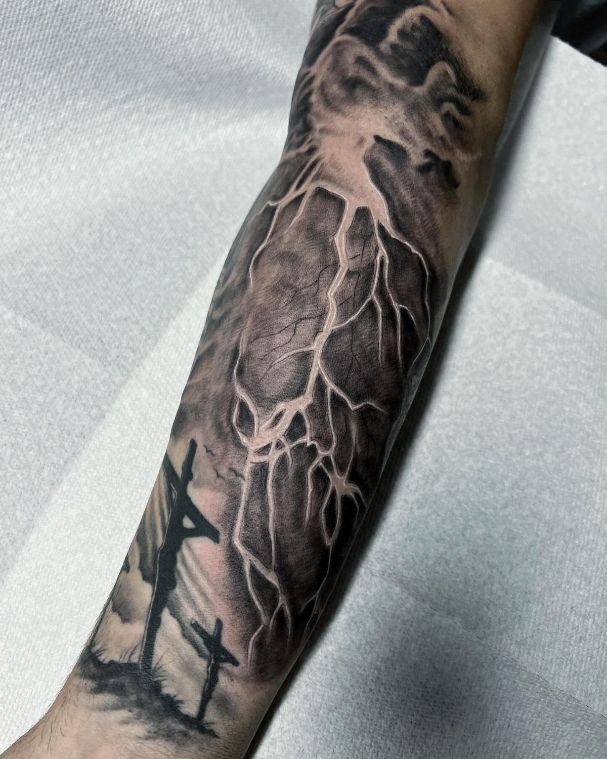 Tattoo on an arm showing dark clouds and lightning bolts with crosses below.