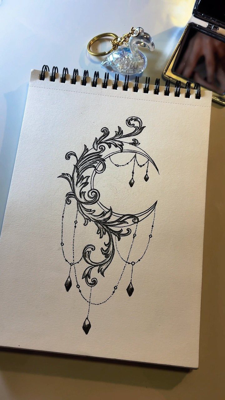 Ornate crescent moon tattoo design with floral swirls and hanging jewels on notebook paper.