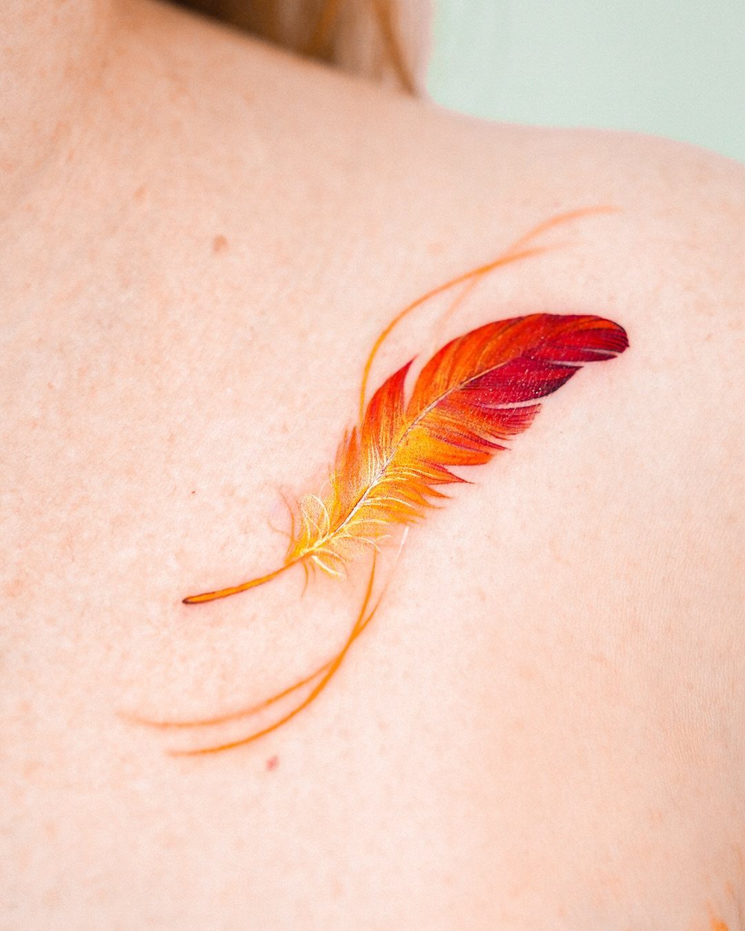 Colorful watercolor feather tattoo in red, orange, and yellow on a shoulder