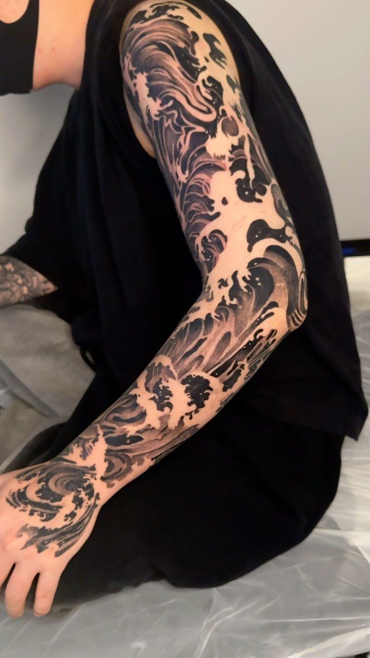 Full sleeve tattoo of crashing ocean waves in black and grey ink.
