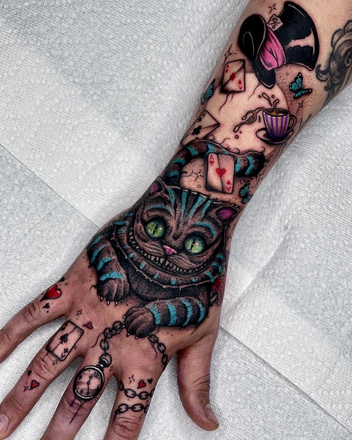 Cheshire Cat tattoo on a hand with playing cards and a Mad Hatter hat.