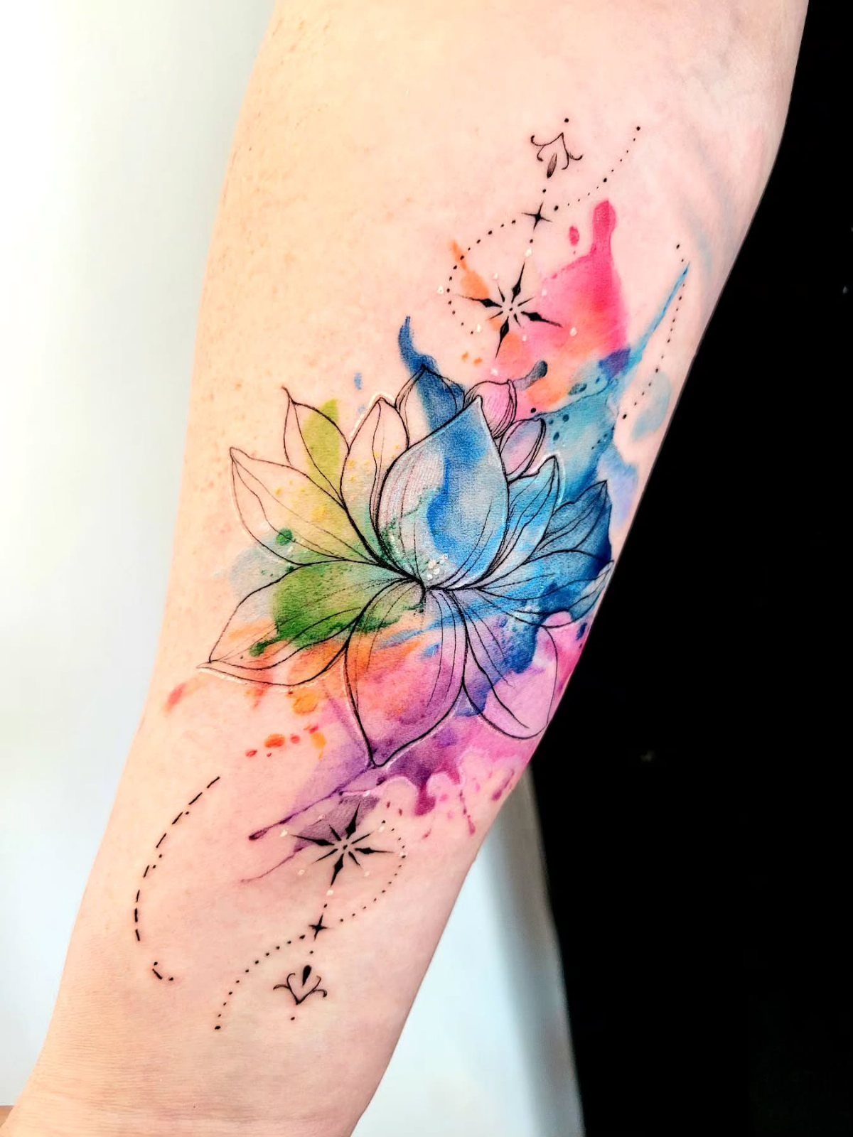 Watercolor lotus flower tattoo on a forearm with vibrant colorful paint splashes.