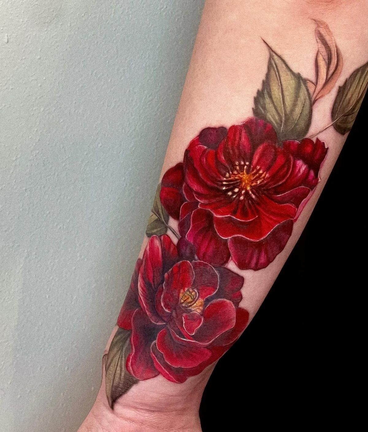 Red peony tattoo with green leaves on a person's forearm