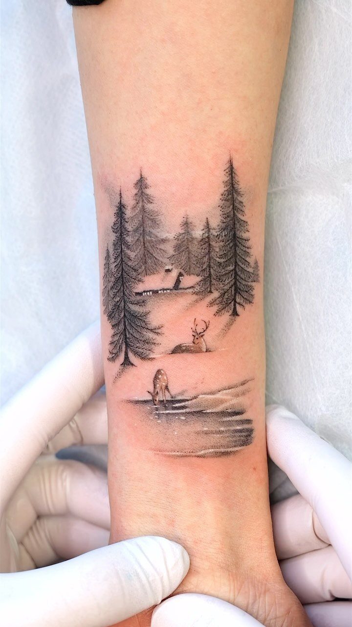 Winter forest tattoo on a wrist featuring pine trees, a cabin, and a deer.