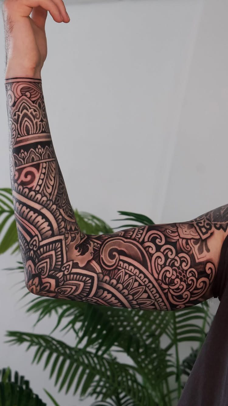 Intricate mandala sleeve tattoo on a person's arm with green foliage in background