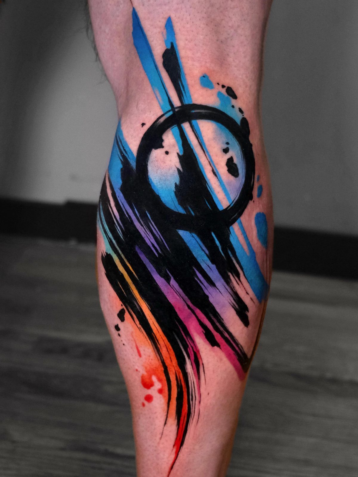 Abstract tattoo on a leg with black brushstrokes and colorful ink splashes.