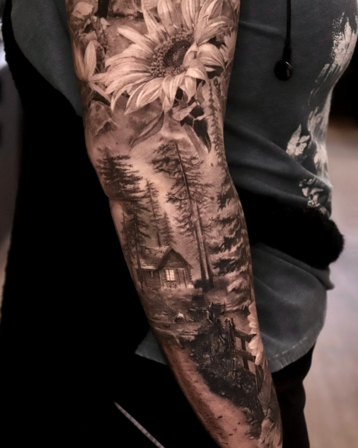 Black and grey arm tattoo featuring a sunflower and a forest landscape with a cabin.