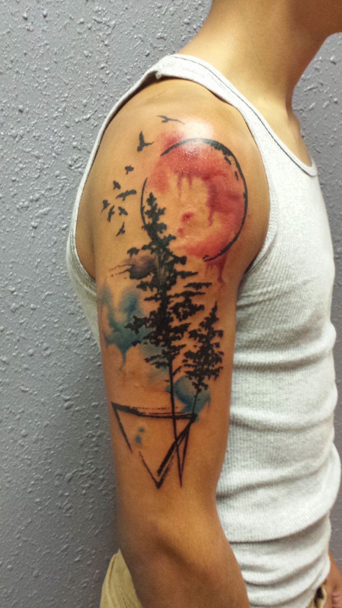 Watercolor tattoo on a man's arm showing pine trees and a red sun.