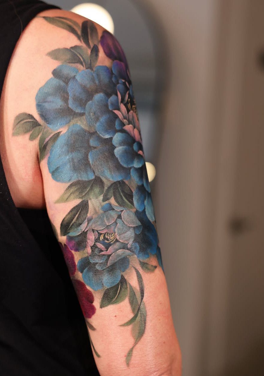 Blue and purple peony tattoo covering the upper arm of a person.