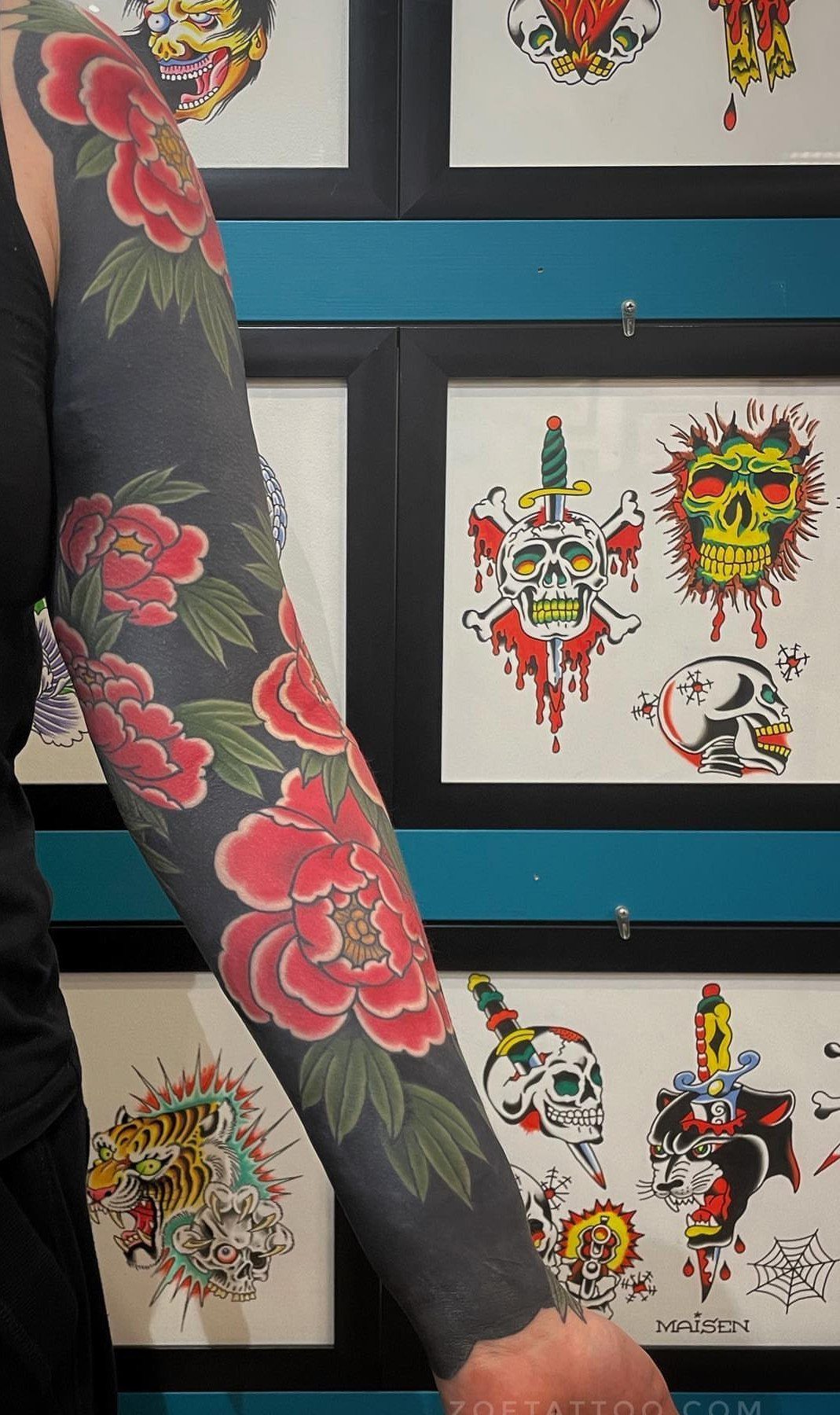 Arm with red peony sleeve tattoo over blackwork against a wall of flash art
