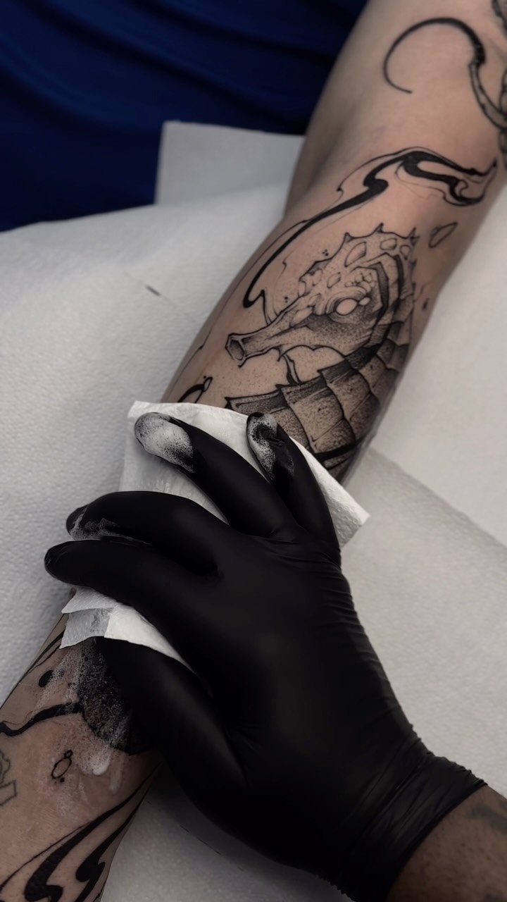 Tattoo artist in black gloves wiping a seahorse tattoo on a person's arm.