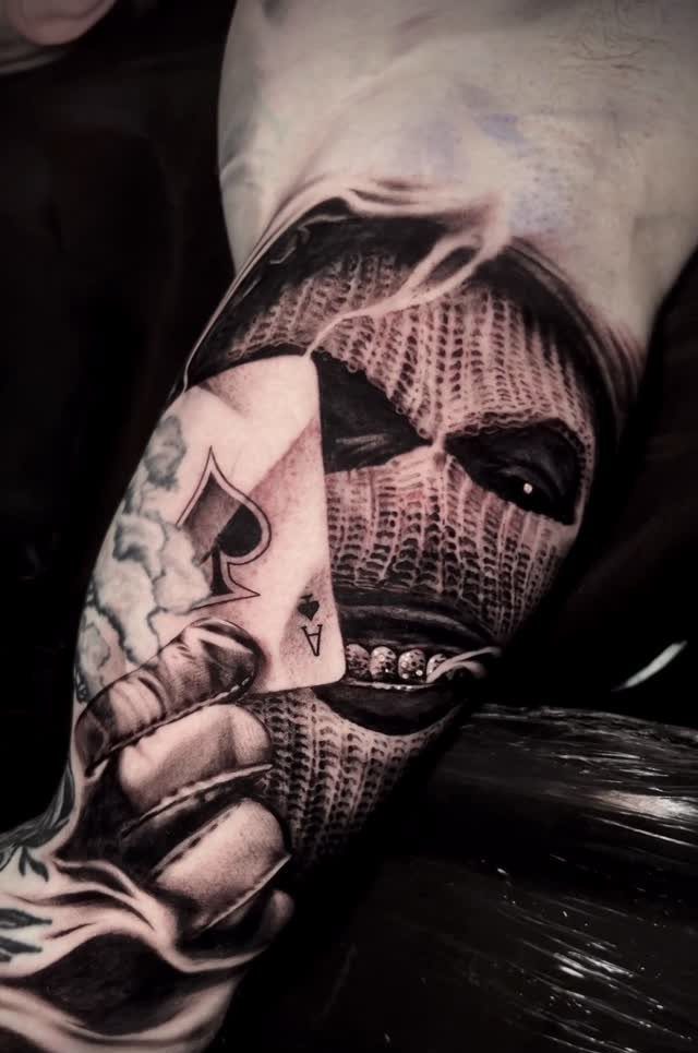 Realism tattoo of a masked figure holding an ace of spades on an arm.