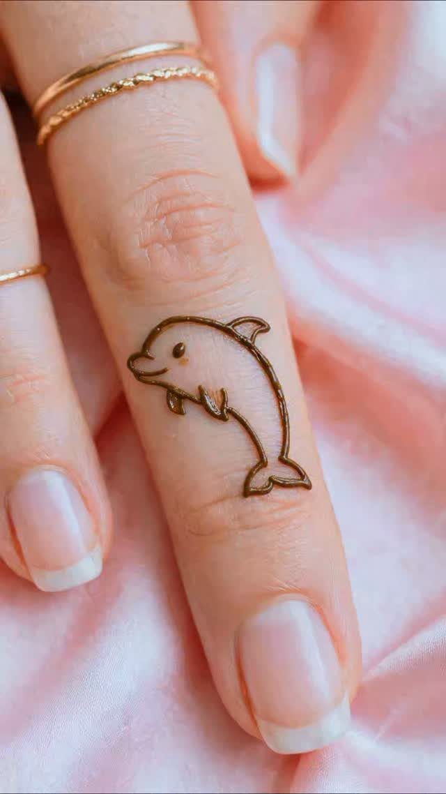 Single dolphin henna tattoo on a finger with gold rings and pink background