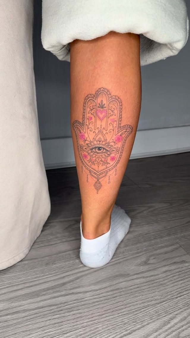 Hamsa hand tattoo with a central eye and pink details on a lower leg.