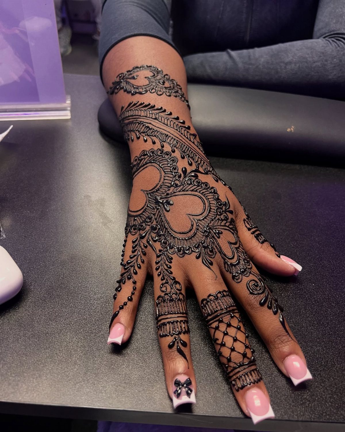 Hand with intricate black henna tattoo featuring a heart and swirly patterns.