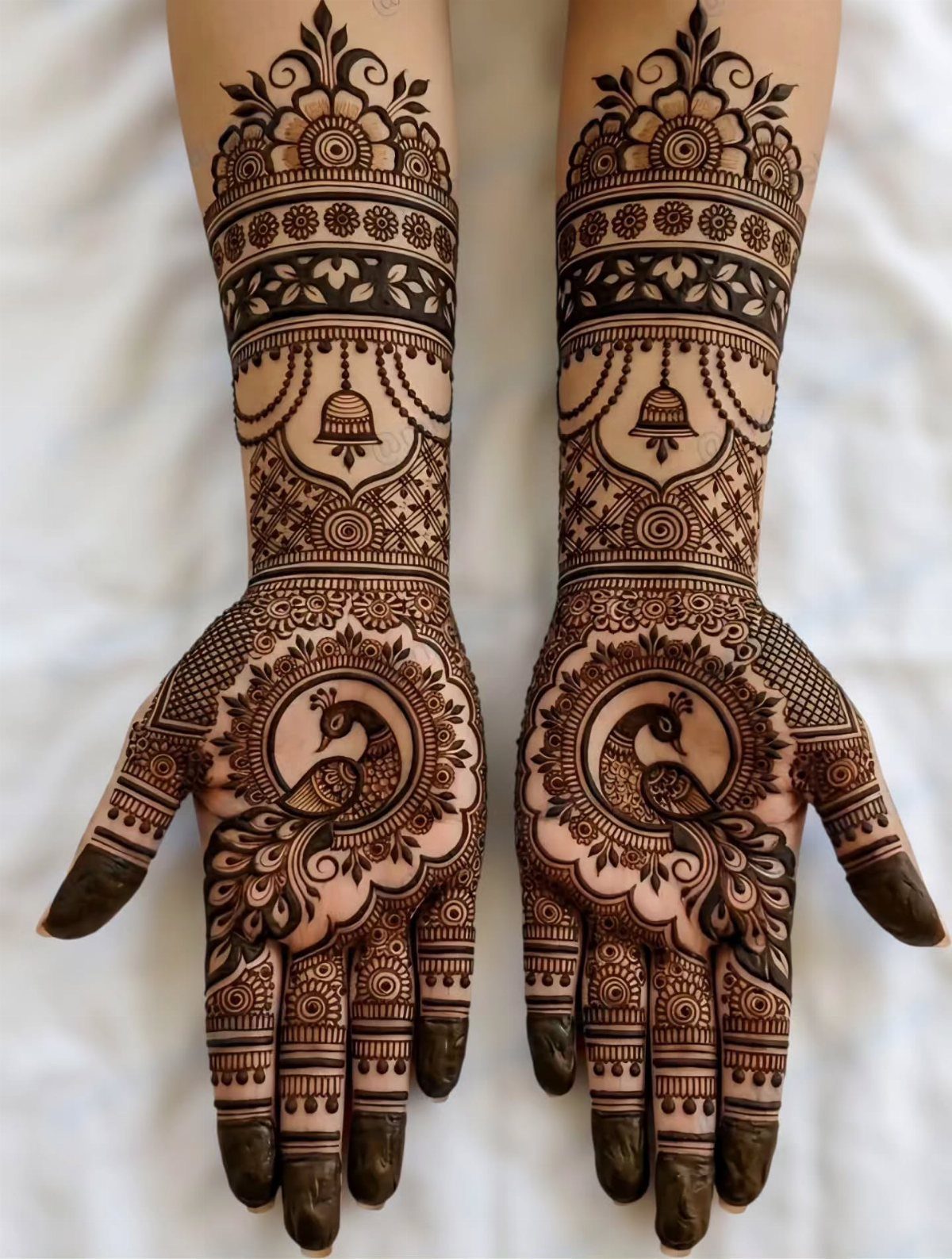 Intricate brown henna designs on two palms and forearms featuring detailed peacocks and florals.
