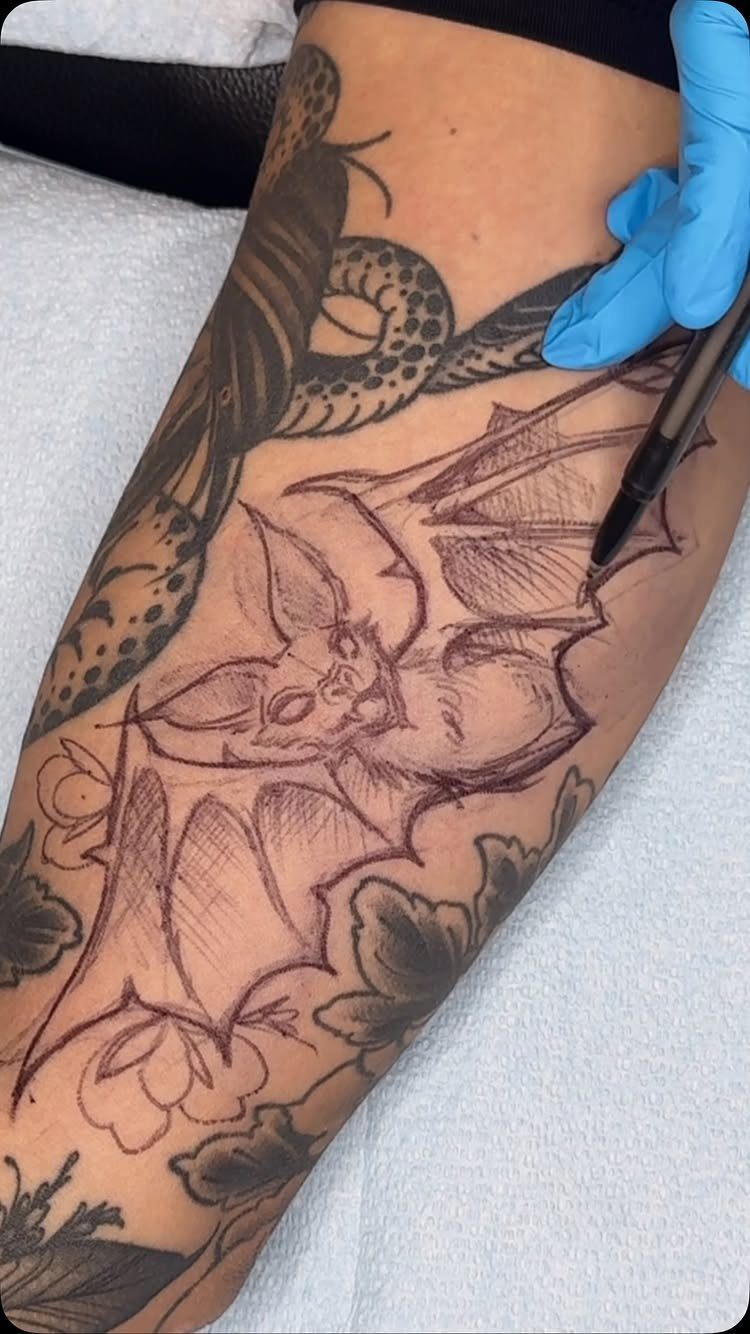 Hand sketching a bat tattoo design on a tattooed arm with a marker