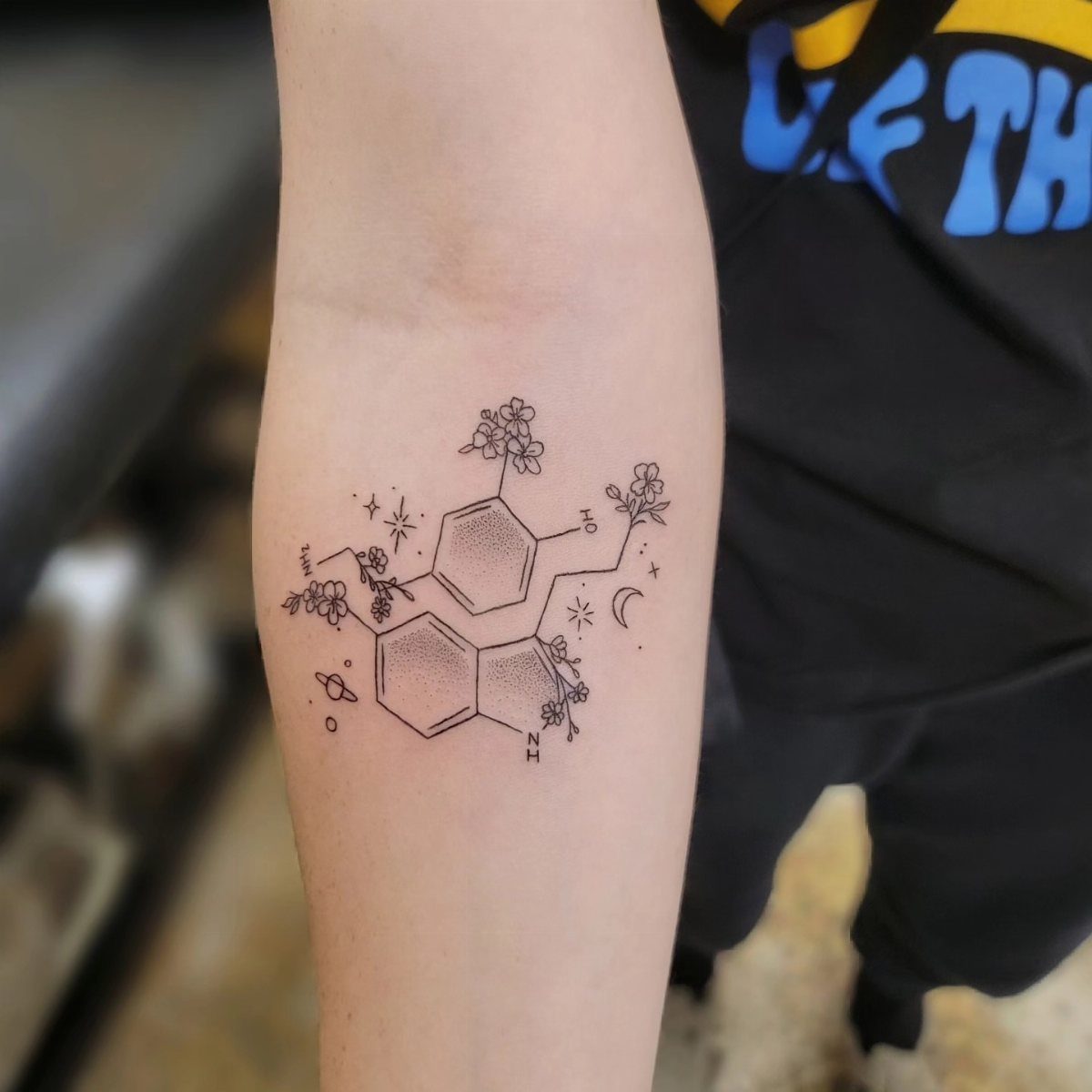 Serotonin molecule tattoo on a forearm decorated with small flowers and celestial elements.