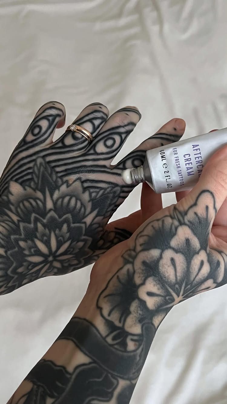 Hands with black tattoos applying aftercare cream from a white tube.