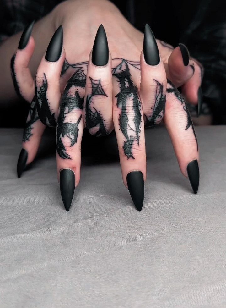 Hand with matte black stiletto nails and detailed black finger tattoos.