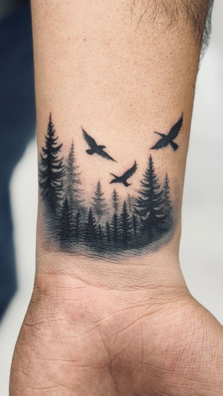 Black ink tattoo of a pine forest and flying birds on a forearm.
