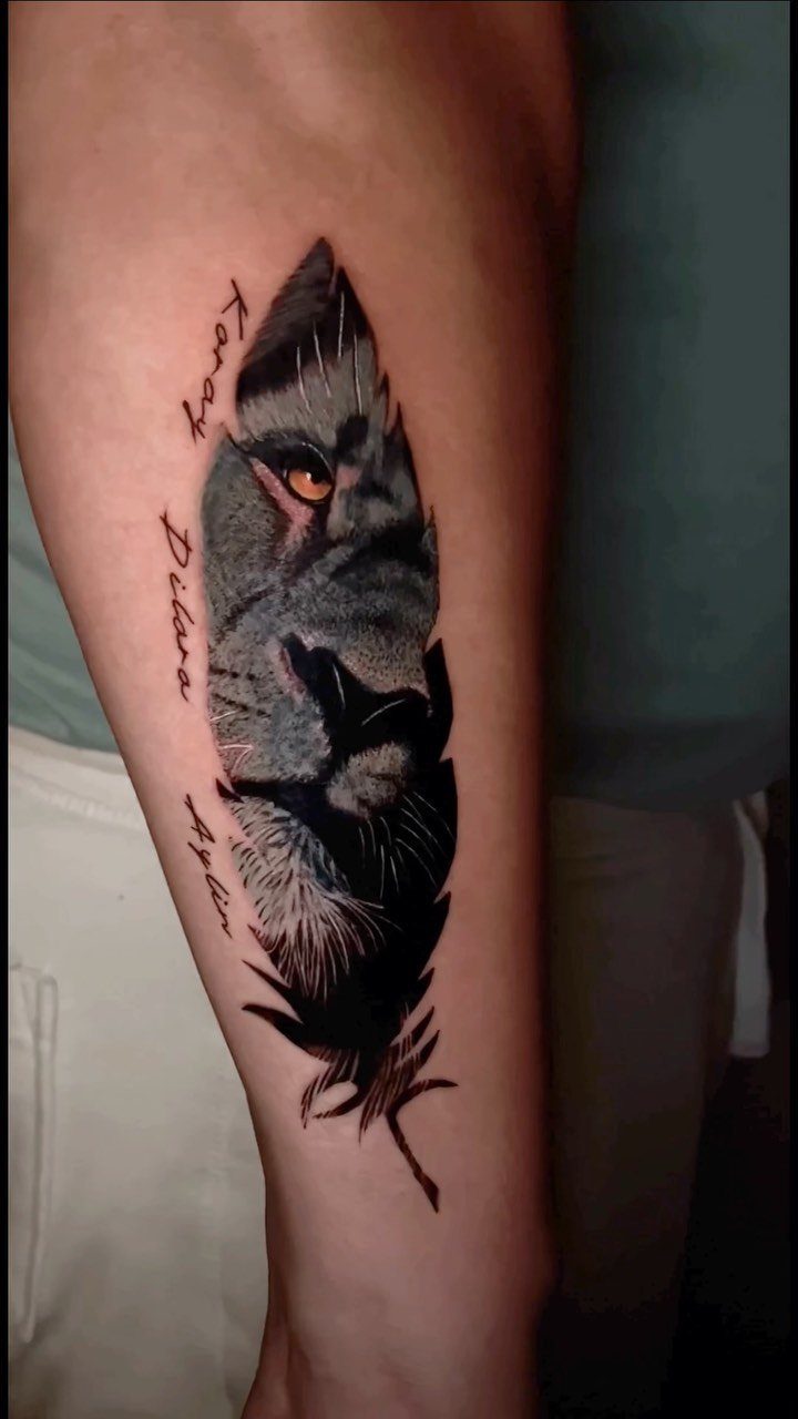Lion face tattoo on a forearm with detailed orange eye and cursive text.