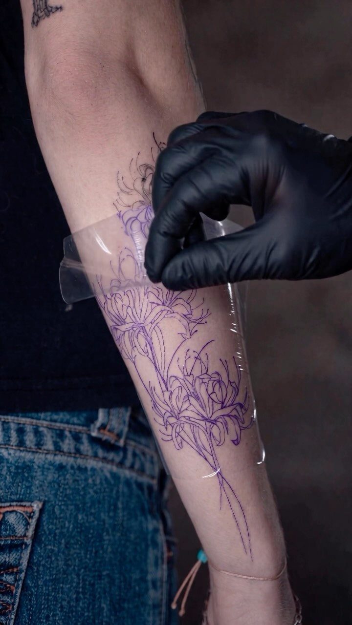 Purple floral tattoo stencil being applied to a person's forearm by gloved hand