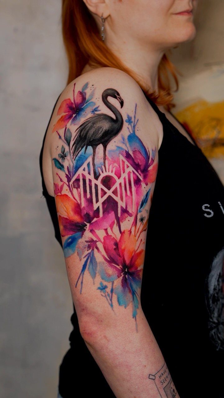 Watercolor tattoo of a black flamingo and colorful flowers on a person's arm.