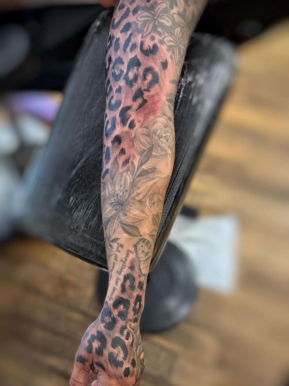 Tattooed arm featuring leopard print and floral designs resting on a black cushion.